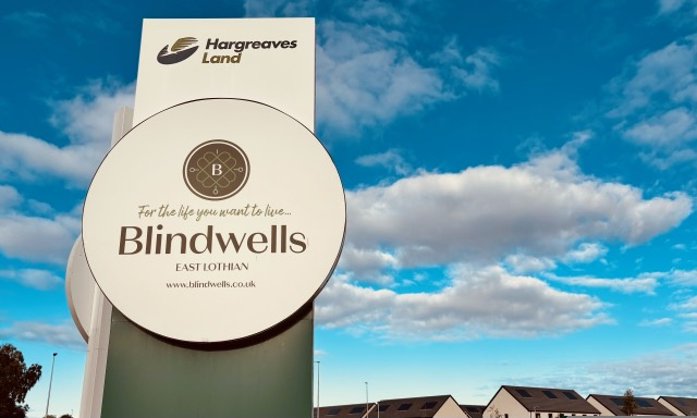 Round Blindwells entrance sign against a blue sky with new homes behind